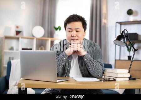 Nachdenklicher asiatischer Mann im karierten Hemd, der zu Hause am Tisch sitzt und auf den Computerbildschirm schaut. Junge Freiberufler über Idee oder die Entscheidung über Arbeitsmomente. Stockfoto