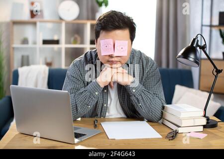 Müde asiatische Geschäftsmann schlafen am Arbeitsplatz mit gefälschten Augen auf Papier Aufkleber gemalt bedeckt seine Augen. Junger Mann sitzt am Tisch mit Laptop und lehnt sich an die Hände. Überarbeiteter Begriff. Stockfoto