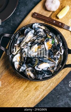 Muscheln in einer cremigen Knoblauchsauce in einer schwarzen Metallplatte auf einem Holzbrett. Stockfoto