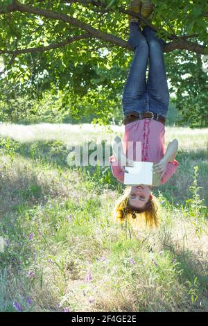 Fröhliches Mädchen hängen auf dem Baum auf den Kopf und Buch lesen Stockfoto