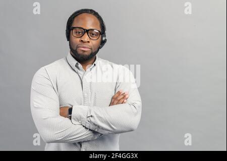 Gutaussehende selbstbewusste gebräunte männliche Bediener und Headset Blick auf die Kamera, mit Armen vor grauem Hintergrund gefaltet stehen, mit Kopieplatz, stolz auf das, was er wirbt, Kundendienstmitarbeiter Porträt Stockfoto