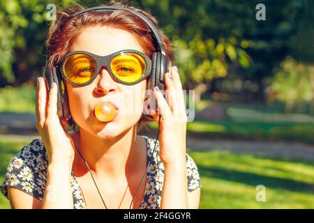 Lustige Mädchen mit Kopfhörern Musik hören und bläst ein Kaugummi. Sorglose junge Frau im lässigen Stil und gelbe Sonnenbrille im Freien im sonnigen su Stockfoto