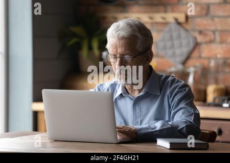 Ein alter männlicher Remote-Mitarbeiter sitzt am Küchentisch mit dem pc Stockfoto