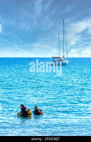 La Herradura, Spanien; 17. Oktober 2020: Zwei Männer in Neoprenanzügen kommen aus dem Meer mit einem schönen Segelboot im Hintergrund Stockfoto