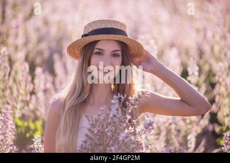 Junge blonde Frau in Strohhut auf dem Hintergrund blühenden Feld von rosa Salbei. Close-up-Porträt schöne Mädchen hält Strauß Blumen in den Händen. W Stockfoto