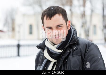 Schöner Mann trägt warme Kleidung im Freien an verschneiten Tag. Winterurlaub. Junger Mann im warmen Mantel lächelt vor der Kamera auf verschwommenem Hintergrund des verschneiten Parks