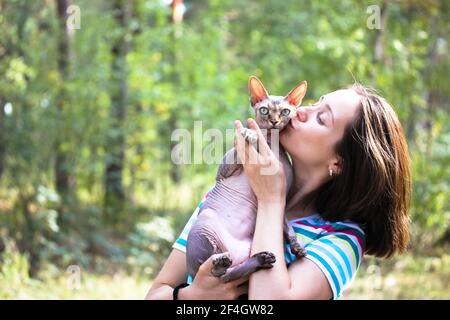 Porträt einer jungen Brünette mit einer kanadischen Sphynx-Katze in einem sommerlichen Wald, hält sie in den Armen, umarmt und küsst sie. Geliebte Glatze. A CO Stockfoto