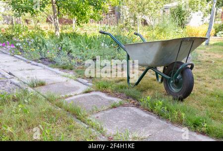 Ein Gartenwagen steht auf einer grünen Wiese in der Nähe eines alten Betonweges vor dem Hintergrund eines Sommergartens in Unschärfe. Stockfoto