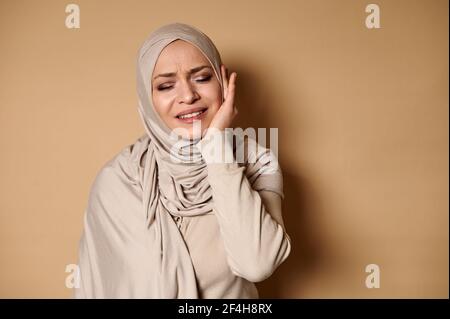 Eine muslimische Frau in einem strengen formellen traditionellen Kleid mit einem Kopf bedeckt mit einem Hijab leidet unter Zahnschmerzen. Beigefarbener Hintergrund mit Kopierbereich Stockfoto