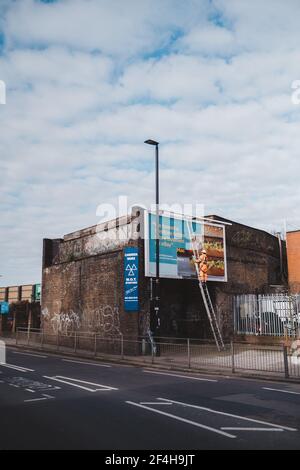 Brentford, West London - 2021,03.21: Arbeiter bereitet Plakatwand für die Installation neuer Werbung vor. Industrieller Kletterer, der auf der Leiter arbeitet - die Unterbringung Stockfoto