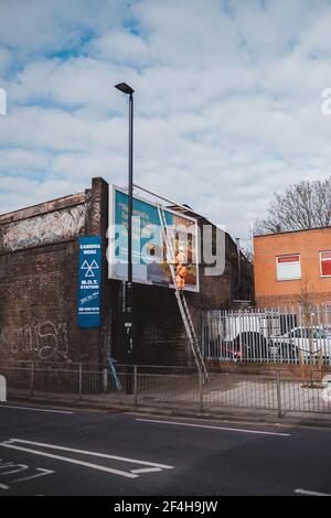 Brentford, West London - 2021,03.21: Arbeiter bereitet Plakatwand für die Installation neuer Werbung vor. Industrieller Kletterer, der auf der Leiter arbeitet - die Unterbringung Stockfoto