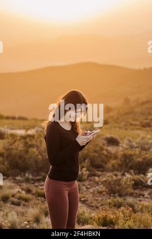 Seitenansicht der entzückten weiblichen Surfen Handy während des Stehens Auf Hügel gegen Berg unter Sonnenuntergang Himmel Stockfoto