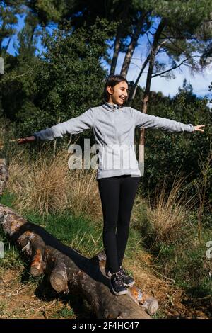 Ganzer Körper von glücklichen jungen Weibchen in Sportbekleidung balancieren auf Gefallener Baumstamm beim Wandern durch grünen Wald bei Sonnenschein Tag Stockfoto