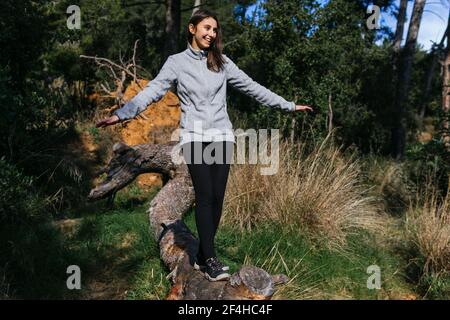 Ganzer Körper von glücklichen jungen Weibchen in Sportbekleidung balancieren auf Gefallener Baumstamm beim Wandern durch grünen Wald bei Sonnenschein Tag Stockfoto