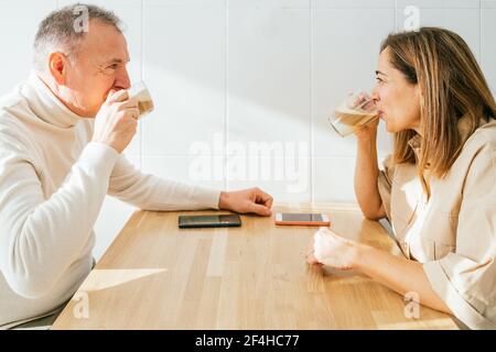 Seitenansicht des Inhalts Erwachsener Paar sitzt am Tisch in Küche und genießen aromatischen Kaffee beim Frühstück und suchen Aufeinander zu Stockfoto