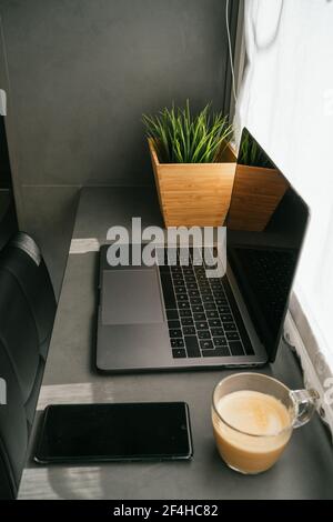 Moderne Laptop und Smartphone auf der Theke mit Tasse platziert Morgenkaffee in der Küche, beleuchtet von Sonnenlicht Stockfoto