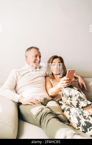 Ein lächelndes Paar mittleren Alters kuschelt auf dem Sofa im Wohnzimmer Und surfen auf dem Smartphone, während Sie sich am Wochenende zu Hause entspannen Stockfoto