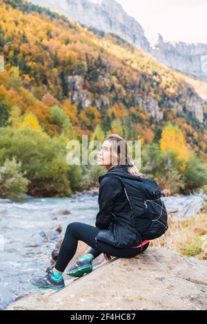 Seitenansicht einer Wandererin mit Rucksack auf einem Felsbrocken In der Nähe des Flusses in den Pyrenäen während des Urlaubs in Ordesa Y Monte Perdido National Stockfoto