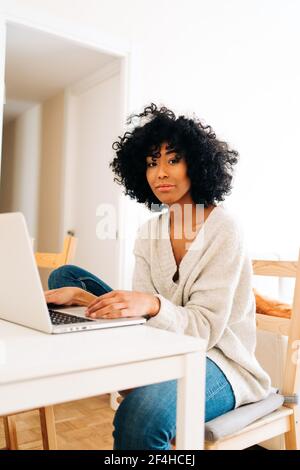 Entzückte afroamerikanische Freiberuflerin, die mit einem Laptop am Tisch sitzt Und die Kamera während der Arbeit an entfernten Standorten im Home Office betrachten Stockfoto