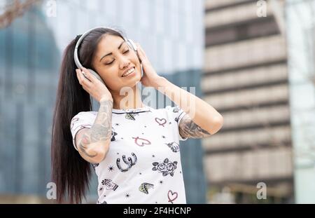 Entzückende junge asiatische Frau in Kopfhörern lächelnd und hörend Musik mit geschlossenen Augen auf verschwommenem Hintergrund der Stadtstraße Stockfoto
