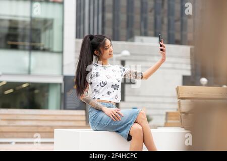 Stilvolle asiatische Frau mit tätowierten Arm auf Bank und ruhen Selfie auf der Stadtstraße Stockfoto