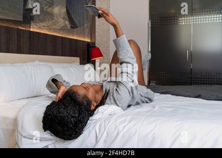 Fröhliche schwarze weibliche Teenager in warmen Pullover und Socken liegen Auf einem bequemen Bett und lachte, während man einen Videoanruf anhatte Smartphone zu Hause Stockfoto