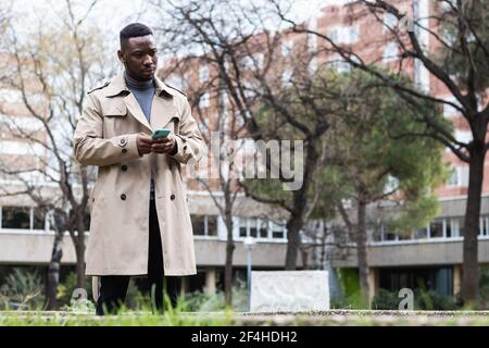 Ernst nachdenklich Erwachsene afroamerikanische Männchen in stilvollem Mantel mit Mobiltelefon in den Händen, das wegschaut, während es auf der Stadt steht Straße im Frühling Tag Stockfoto