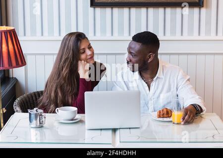 Glückliches multirassisches Paar sitzt am Tisch und browsen Laptop zusammen Beim Frühstück im stilvollen Zimmer zu Hause Stockfoto