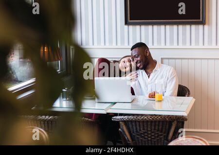 Glückliches multirassisches Paar, das zusammen Laptop durchstöbert, während es am Tisch sitzt Im stilvollen Zimmer zu Hause Stockfoto