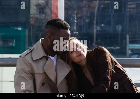 Stilvolle liebevolle multiethnische Paar in eleganten Outfits während des Sitzens Bank am Busbahnhof in der Stadt Stockfoto