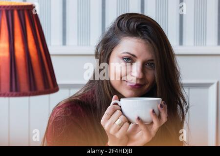 Verträumte Erwachsene Frau mit Tasse heißen Kaffee in den Händen Blick auf die Kamera und lächeln, während Sie Freizeit in genießen Café Stockfoto