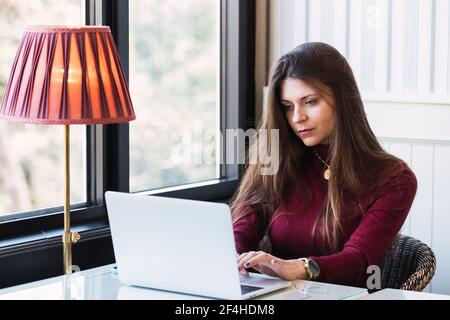 Serious junge freiberufliche Freiberuflerin sitzt am Tisch neben Fenster und Mit einem Laptop arbeiten, während Sie im gemütlichen Café aus der Ferne arbeiten Stockfoto