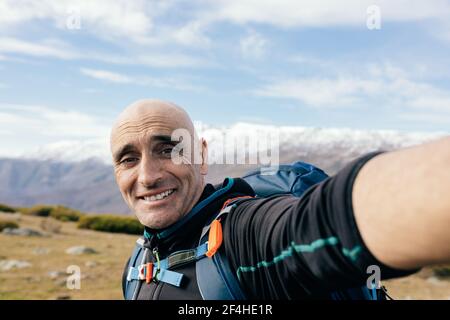 Fröhliche Erwachsene männliche Kletterer in aktivewear Blick auf Kamera, während Selfie gegen majestätische schneebedeckte Felsspitzen an sonnigen Tagen Stockfoto