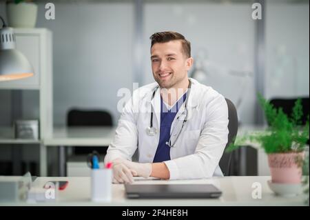 Ein netter junger Arzt sitzt mit an seinem Schreibtisch Ein Stethoskop und Handschuhe an Stockfoto