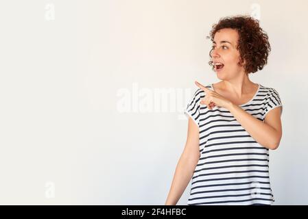 Lockig behaart junge kaukasische Frau mit überrascht Gesicht auf grau Hintergrund isoliert Zeigefinger zur Seite mit viel Kopie Platz Stockfoto