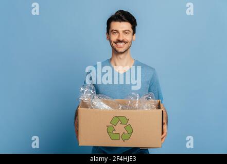 Lächelnder unrasiert gewissenhafter junger Mann in blauem T-Shirt hält einen Karton voll Plastikflaschen zum Recycling, lächelt in die Kamera, steht auf isoliertem blauem Hintergrund, umweltfreundliches Konzept Stockfoto