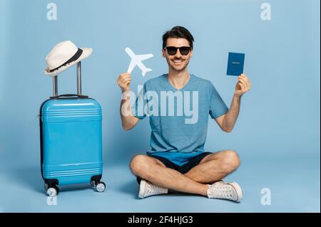 Urlaubszeit. Fröhlicher kaukasischer männlicher Tourist in Sonnenbrillen und Sommerkleidung sitzt auf isoliertem blauem Hintergrund, hält Pass in der einen Hand, Flugzeug in der anderen, blauer Koffer steht in der Nähe und lächelt Stockfoto
