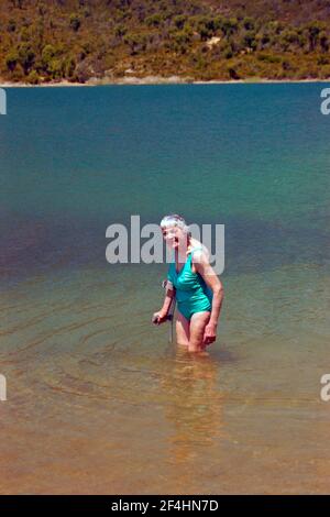 Lächelnde ältere Frau im Badeanzug, knietief im türkisfarbenen See stehend, sich auf einer Krücke feststehend. Stockfoto