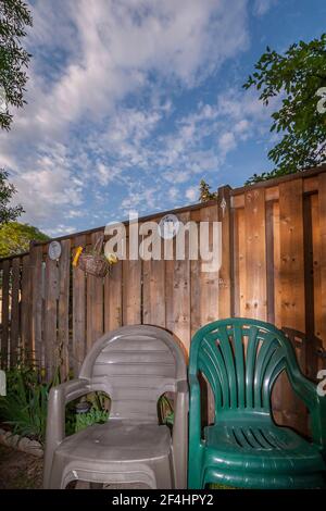 Ein grauer und ein grüner Plastikstuhl gegen einen Holzstuhl Zaun und ein dramatischer Himmel im Hintergrund Stockfoto
