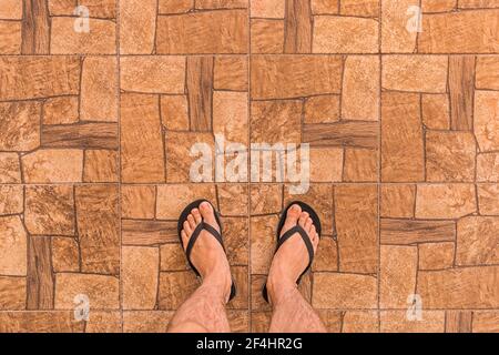 Die Beine eines Mannes in schwarzen Flip Flops stehen auf einem braunen Fliesenboden mit abstraktem Steinmuster im Hintergrund, Draufsicht. Stockfoto