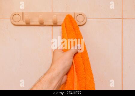 Die Hand des Mannes nimmt ein orangefarbenes Handtuch im Badezimmer. Pflege- und Hygienekonzept. Stockfoto