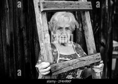 Eine alte Frau schaut hinter einer Leiter im Dorf heraus. Schwarzweiß-Foto. Stockfoto