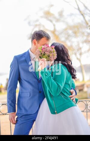 Brautpaar-Porträt einer Braut in einer grünen Jacke und eines Bräutigams in einem blauen Anzug mit einem Blumenstrauß in der Mitte an einem sonnigen Frühlingsnachmittag Stockfoto