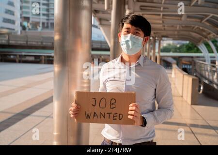 Junge aufgeregt asiatischen Mann trägt covid-19 Schutz Gesichtsmaske fragen Für Job über Nachricht auf dem Schild nach Verlust des Jobs fällig Zu Coronavirus Pandemie und Ausbruch Stockfoto