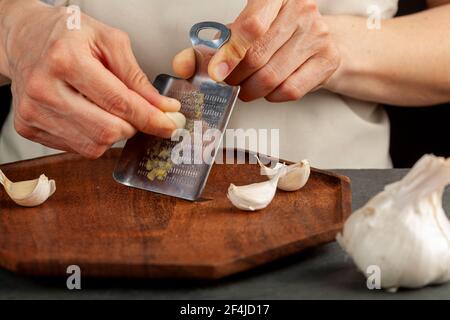 Hände einer kaukasischen Frau, die Knoblauchzehen mit schaufelförmigen Mini-Reibe aus Metall auf einem Holzteller reibt. Dieses kleine Stück kann auch als Ingwer g verwendet werden Stockfoto