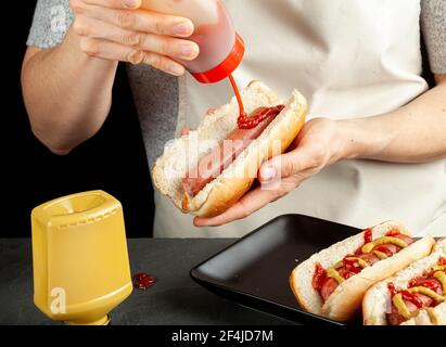 Eine kaukasische Frau bereitet Hot Dog Sandwiches zu. Sie quetscht Tomatenketchup aus der Plastikflasche auf eine Rinderwurst in einem Hot Dog Brötchen. Oth Stockfoto