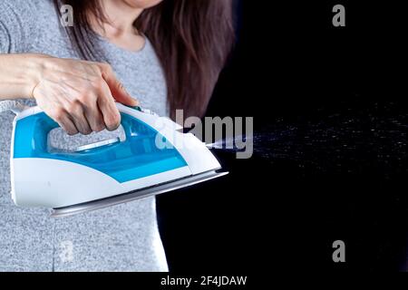 Eine Frau hält ein Dampfbügeleisen in der Hand, das aus einem winzigen Loch davor druckfestes Wasser austritt. Wasser spritzen kann gegen dunkle b gesehen werden Stockfoto