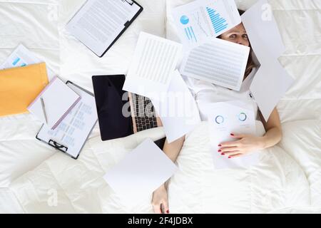 Müde Frau liegt auf dem Bett mit Laptop und Business-Charts Stockfoto