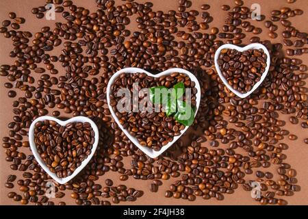 Geröstete braune Kaffeebohnen auf braunem Hintergrund. Draufsicht. Herzförmige Kaffeebohnen in einer weißen Schale. Stockfoto