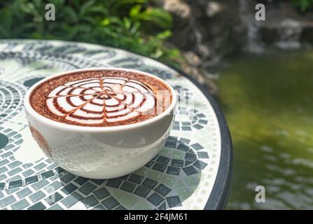 Schickes Design auf heißen Kaffee in weißen heißen Kaffeebecher, auf einem Keramik-Tisch platziert. Die Kulisse eines Wasserfalls, üppiges Grün und Dampf schwebte nach ma Stockfoto